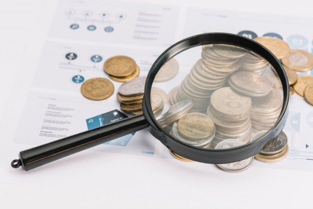 moedas sobre conta com uma lupa por cima para exemplificar Quais impostos pagamos