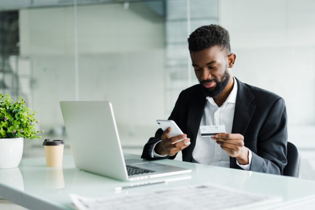 homem empresário com um cartão de crédito para pessoa jurídica na mão em frente ao laptop e com o celular na outra mão