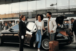 Casal sorridente de brasileiros desembarcando de um carro de luxo em frente ao terminal de aeroporto, representando os top 3 cartões que oferecem transfer com serviço de motorista particular.
